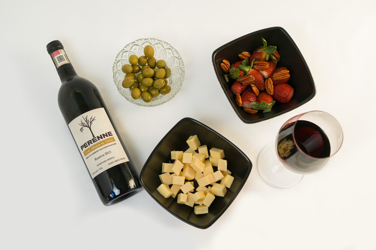 Top view of red wine bottle with two glasses and three plates featuring strawberries, olives, and cheese on white background