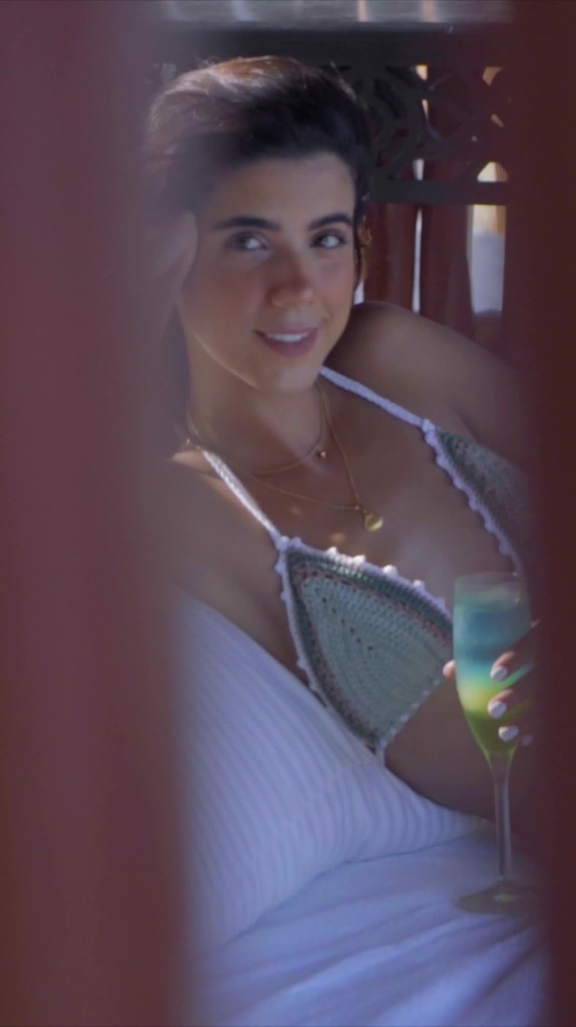 Regina Ayné modeling a handwoven bikini while playfully interacting with the camera in a beach setting, with natural light and ocean scenery.
