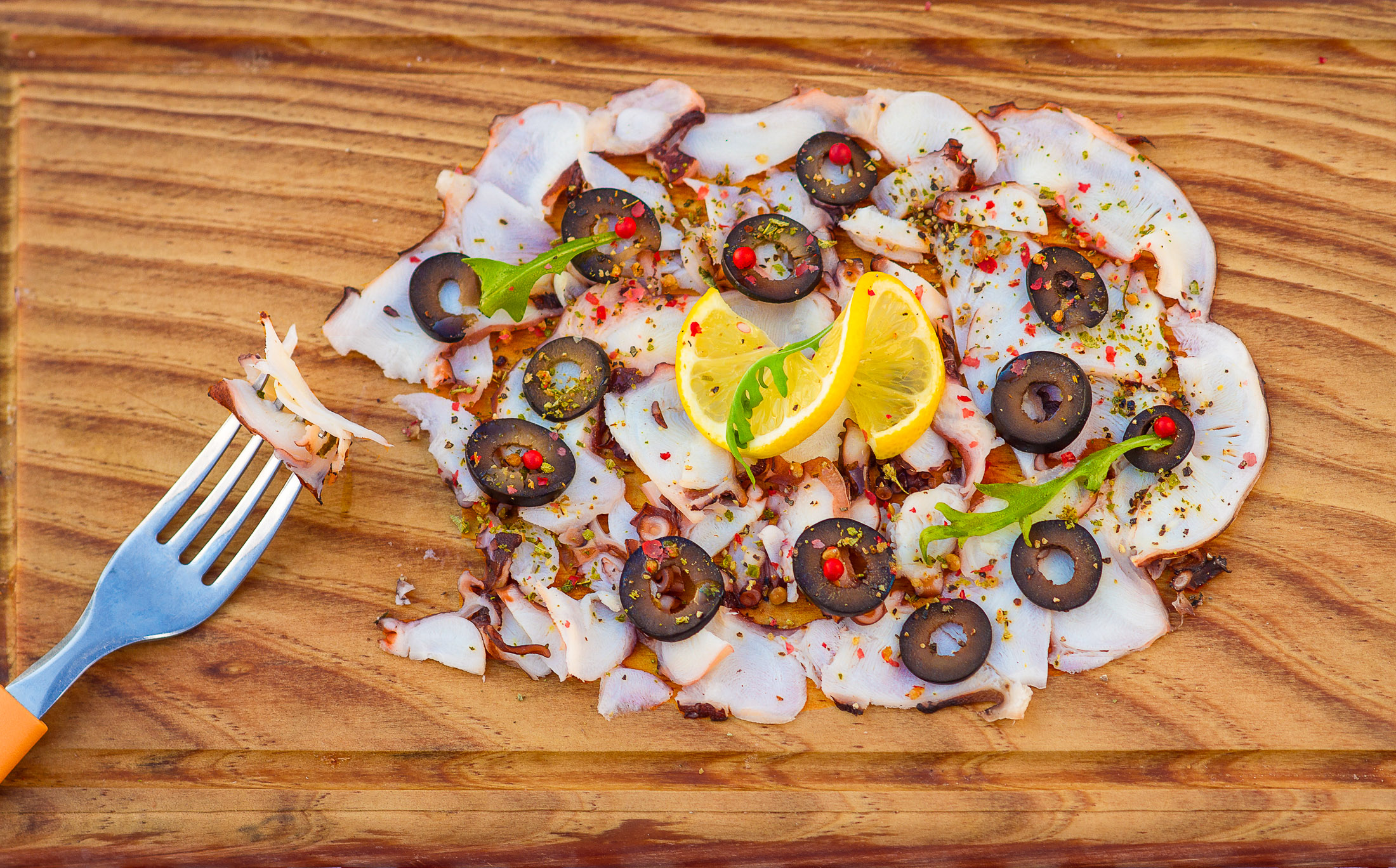 Top view of octopus carpaccio on wooden board with fork bite, black olives, red pepper, and fresh lemon outdoors