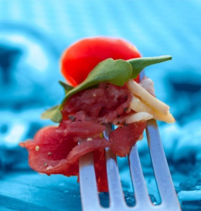 food photography - A bite ready to go into your mouth, a fork with beef carpaccio, a green leaf, and half a cherry tomato